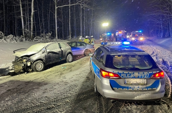 miejsce wypadku, na drodze stoi radiowóz policyjny, na poboczu drogi stoją pojazdy osobowe uczestniczące w zdarzeniu, dalej pojazdy służb ratunkowych i strażacy.