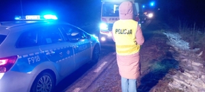 policjantka na miejscu zdarzenia drogowego, ubrana po cywilnemu, ma założoną kamizelkę z napisem policja. na drodze stoi radiowóz policyjny, dalej karetka pogotowia.