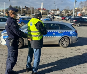 policjantka zakłada pieszemu kamizelkę odblaskową, dalej widoczny radiowóz oraz parking samochodowy i ulica.