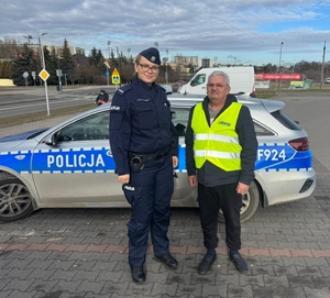 policjantka zakłada pieszemu kamizelkę odblaskową, dalej widoczny radiowóz oraz parking samochodowy i ulica.