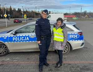 policjantka założyła pieszejkamizelkę odblaskową, dalej widoczny radiowóz oraz parking samochodowy i ulica.