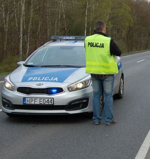policjant po cywilnemu ma założoną kamizelką z napisem policja, stoi na drodze przy radiowozie policyjnym.