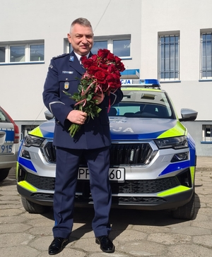 policjant trzyma kwiaty w dłoniach, w tle radiowóz policyjny oraz budynek komendy.