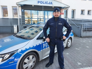 umundurowany policjant stoi przy radiowozie, w tle budynek i wejście główne do budynku komendy policji w Bełchatowie.