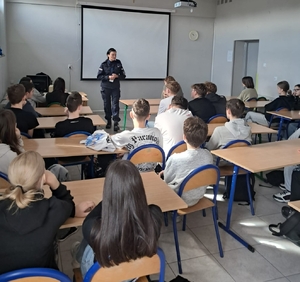 policjantka na spotkaniu z młodzieżą w szkole, uczniowie siedzą przy ławkach a policjantka prowadzi prelekcję.