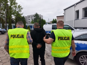 policjanci ubrani po cywilnemu, ubrani w kamizelki odblaskowe z napisem policja prowadzą zatrzymanego mężczyznę. Zdjęcie zrobione na placu wewnętrznym komendy policji, w tle radiowozy policyjne i budynek garaży.