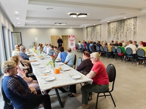 policjant na spotkaniu z seniorami, wszyscy siedzą na sali przy stolach, policjant prowadzi prelekcję.