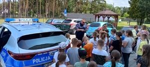 policjant n spotkaniu z grupą dzieci , stoją wszyscy przy radiowozie, policjant rozmawia z uczniami.