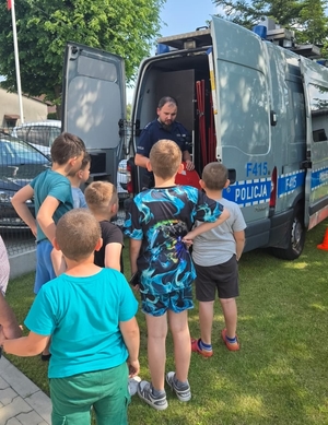 policjant n spotkaniu z grupą dzieci , stoją wszyscy przy radiowozie, policjant rozmawia z uczniami.