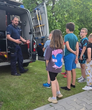 policjant n spotkaniu z grupą dzieci, stoją wszyscy przy radiowozie, policjant rozmawia z dziećmi na spotkaniu w plenerze.