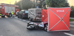 miejsce wypadku drogowego. na drodze osobówka z przyczepką, obok na drodze za przyczepką leży motocykl. Rozstawiony jest również parawan czerwony, dalej widać policjanta, który wykonuje oględziny miejsca zdarzenia. a także radiowóz straży pożarnej.