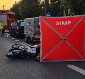miejsce wypadku drogowego. na drodze osobówka z przyczepką, obok na drodze za przyczepką leży motocykl. Rozstawiony jest również parawan czerwony, dalej widać policjanta, który wykonuje oględziny miejsca zdarzenia. a także radiowóz straży pożarnej.