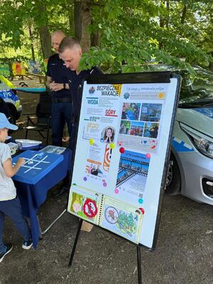 policjanci na pikniku rodzinnym CZAS w LAS, organizowanym przez Nadleśnictwo Bełchatów. Prowadza działania profilaktyczne związane z bezpieczeństwem w czasie wakacji. Stoisko policyjne odwiedzają dzieci i dorośli. Na stoliku znajdują się materiały profilaktyczne dotyczące bezpieczeństwa . W tle radiowozy policyjne i umundurowani policjanci rozmawiający z odwiedzającymi.