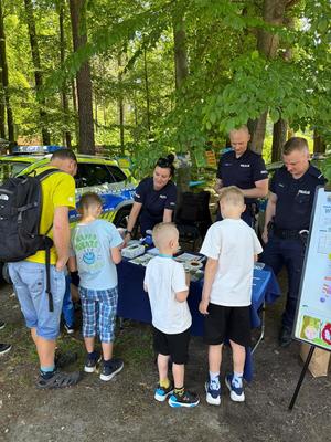policjanci na pikniku rodzinnym CZAS w LAS, organizowanym przez Nadleśnictwo Bełchatów. Prowadza działania profilaktyczne związane z bezpieczeństwem w czasie wakacji. Stoisko policyjne odwiedzają dzieci i dorośli. Na stoliku znajdują się materiały profilaktyczne dotyczące bezpieczeństwa . W tle radiowozy policyjne i umundurowani policjanci rozmawiający z odwiedzającymi.