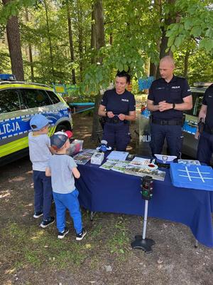 policjanci na pikniku rodzinnym CZAS w LAS, organizowanym przez Nadleśnictwo Bełchatów. Prowadza działania profilaktyczne związane z bezpieczeństwem w czasie wakacji. Stoisko policyjne odwiedzają dzieci i dorośli. Na stoliku znajdują się materiały profilaktyczne dotyczące bezpieczeństwa . W tle radiowozy policyjne i umundurowani policjanci rozmawiający z odwiedzającymi.