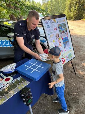 policjanci na pikniku rodzinnym CZAS w LAS, organizowanym przez Nadleśnictwo Bełchatów. Prowadza działania profilaktyczne związane z bezpieczeństwem w czasie wakacji. Stoisko policyjne odwiedzają dzieci i dorośli. Na stoliku znajdują się materiały profilaktyczne dotyczące bezpieczeństwa . W tle radiowozy policyjne i umundurowani policjanci rozmawiający z odwiedzającymi.