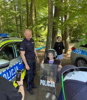 policjanci na pikniku rodzinnym CZAS w LAS, organizowanym przez Nadleśnictwo Bełchatów. Prowadza działania profilaktyczne związane z bezpieczeństwem w czasie wakacji. Stoisko policyjne odwiedzają dzieci i dorośli. Na stoliku znajdują się materiały profilaktyczne dotyczące bezpieczeństwa . W tle radiowozy policyjne i umundurowani policjanci rozmawiający z odwiedzającymi.