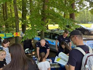 policjanci na pikniku rodzinnym CZAS w LAS, organizowanym przez Nadleśnictwo Bełchatów. Prowadza działania profilaktyczne związane z bezpieczeństwem w czasie wakacji. Stoisko policyjne odwiedzają dzieci i dorośli. Na stoliku znajdują się materiały profilaktyczne dotyczące bezpieczeństwa . W tle radiowozy policyjne i umundurowani policjanci rozmawiający z odwiedzającymi.