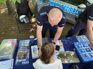 policjanci na pikniku rodzinnym CZAS w LAS, organizowanym przez Nadleśnictwo Bełchatów. Prowadza działania profilaktyczne związane z bezpieczeństwem w czasie wakacji. Stoisko policyjne odwiedzają dzieci i dorośli. Na stoliku znajdują się materiały profilaktyczne dotyczące bezpieczeństwa . W tle radiowozy policyjne i umundurowani policjanci rozmawiający z odwiedzającymi.