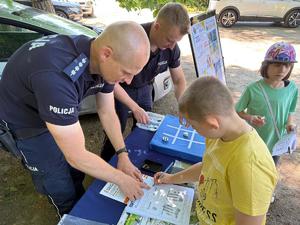 policjanci na pikniku rodzinnym CZAS w LAS, organizowanym przez Nadleśnictwo Bełchatów. Prowadza działania profilaktyczne związane z bezpieczeństwem w czasie wakacji. Stoisko policyjne odwiedzają dzieci i dorośli. Na stoliku znajdują się materiały profilaktyczne dotyczące bezpieczeństwa . W tle radiowozy policyjne i umundurowani policjanci rozmawiający z odwiedzającymi.