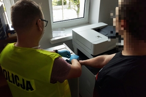 policjanci prowadza zatrzymanego mężczyznę, zdjęcie zrobione na placu wewnętrznym komendy, w tle radiowozy i budynki.