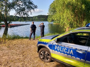 policjanci patrolujący teren nad woda, radiowóz stoi przy zbiorniku wodnym.