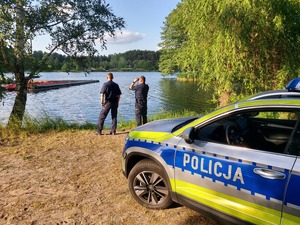 policjanci patrolujący teren nad woda, radiowóz stoi przy zbiorniku wodnym.