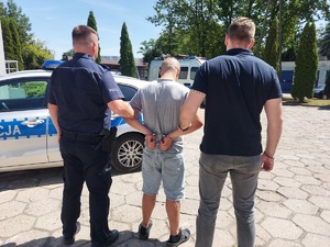 dwóch policjantów, jeden w umundurowaniu służbowym, drugi po cywilnemu prowadzi zatrzymanego mężczyznę, zdjęcie zrobione na placu wewnętrznym komendy policji, obok stoją radiowozy , widać też budynki.