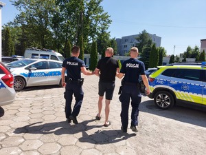 dwóch policjantów prowadzi zatrzymanego mężczyznę, zdjęcie zrobione na placu wewnętrznym komendy policji, obok stoją radiowozy , widać też budynki.