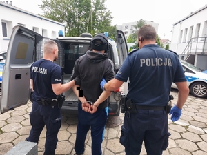 policjanci prowadzą zatrzymanego mężczyznę, zdjęcie zrobione na dziedzińcu komendy, widać też radiowozy policyjne.