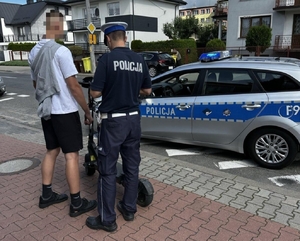 policjant rozmawia z kierową hulajnogi, dalej widoczny radiowóz, droga, miejsce publiczne.