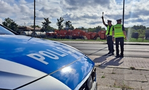 policjanci pełniący służbę w terenie, stoją przy drodze, obok radiowóz. policjant daje sygnał do zatrzymania pojazdu.