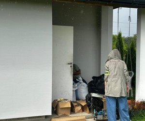 Policjantka z opaską z napisem policja stoi przed domem, na podłodze leżą kartonowe pudełka i worki jutowe.