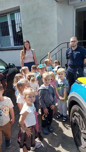 spotkanie policjantów z uczniami. policjant z dziećmi i nauczycielkami stoją przy radiowozie przed budynkiem posterunku policji w Szczercowie.