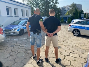 policjant w cywilnych ubraniach prowadzi zatrzymanego mężczyznę. Zdjęcie zrobione na placu wewnętrznym komendy policji, w tle radiowozy policyjne i budynki.