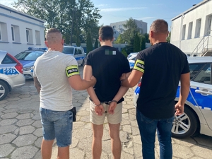 policjanci w cywilnych ubraniach prowadzą zatrzymanego mężczyznę. Zdjęcie zrobione na placu wewnętrznym komendy policji, w tle radiowozy policyjne i budynki.