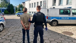 policjant prowadzi zatrzymanego mężczyznę. Zdjęcie zrobione na placu wewnętrznym komendy, w tle radiowozy i budynki.