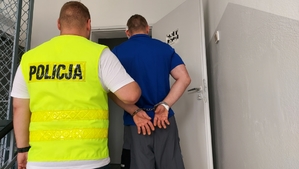 policjant prowadzi zatrzymanego mężczyznę. Zdjęcie zrobione w pomieszczeniu służbowym komendy policji.