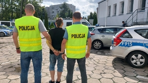 policjanci służby kryminalnej ubrani po cywilnemu z założonymi kamizelkami z napisem policja prowadzą zatrzymanego mężczyznę. Ma on założone kajdanki na ręce trzymane z tylu. Zdjęci pokazuje również plac wewnętrzny komendy policji, widoczne są radiowozy policyjne oraz budynki.