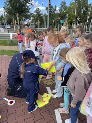 policjant pokazuje chłopcu na czym polega praca policjanta ruchu drogowego w obrębie przejścia dl pieszych, policjanci uczą dzieci prawidłowych zachowań na przejściu dla pieszych. policjanci rozdają dzieciom kamizelki odblaskowe.