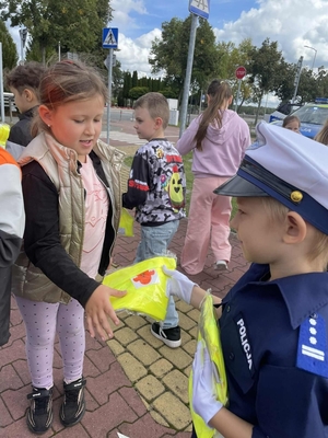 na zdjęciu dzieci, chłopiec w przepraniu &quot;policjanta&quot;, dzieci trzymają kamizelki odblaskowe.