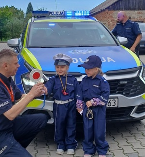 policjant pokazuje chłopcom tarczę sygnalizacyjną. dalej widoczny radiowóz.