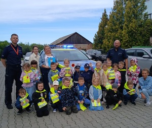 dwóch policjantów razem z grupą przedszkolaków w tle radiowóz. Zdjęcie zrobione na dziedzińcu posterunku.