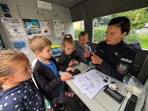 policjantka na spotkaniu z uczniami, dzieci siedzą w radiowozie profilaktycznym, policjantka pokazuje im na czym polega daktyloskopia.