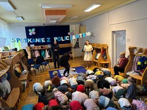 Policjantka stoi przed siedzącymi na podłodze dziećmi w przedszkolu.