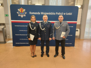 Komendant wraz z policjantką i policjantem stoją na tle baneru Komendy Wojewódzkiej Policji w Łodzi.