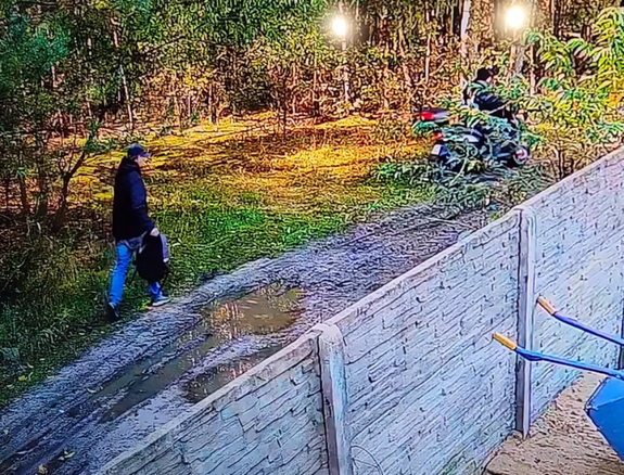 Mężczyzna idzie za jadącym mężczyzną na skuterze za ogrodzeniem.