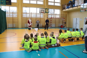 Komendant wraz z wójtem i mikołajem stoją przed dziećmi ubranymi w kamizelki odblaskowe.