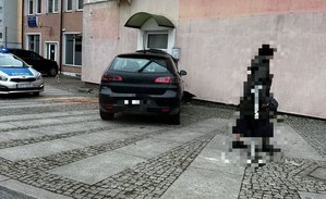 Rozbity samochód przy uszkodzonym budynku, w tle radiowóz.