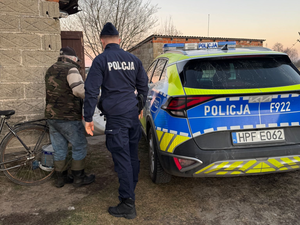 Policjant przy radiowozie, mężczyzna z materacem idzie w kierunku domu.